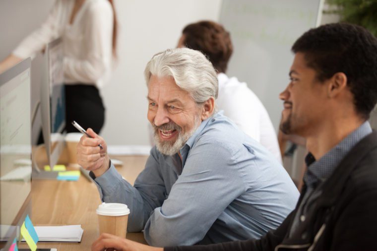 Smiling senior employee discussing email with african colleague at workplace, happy older worker talking to black coworker joking about online computer work, aged and young managers laugh in office